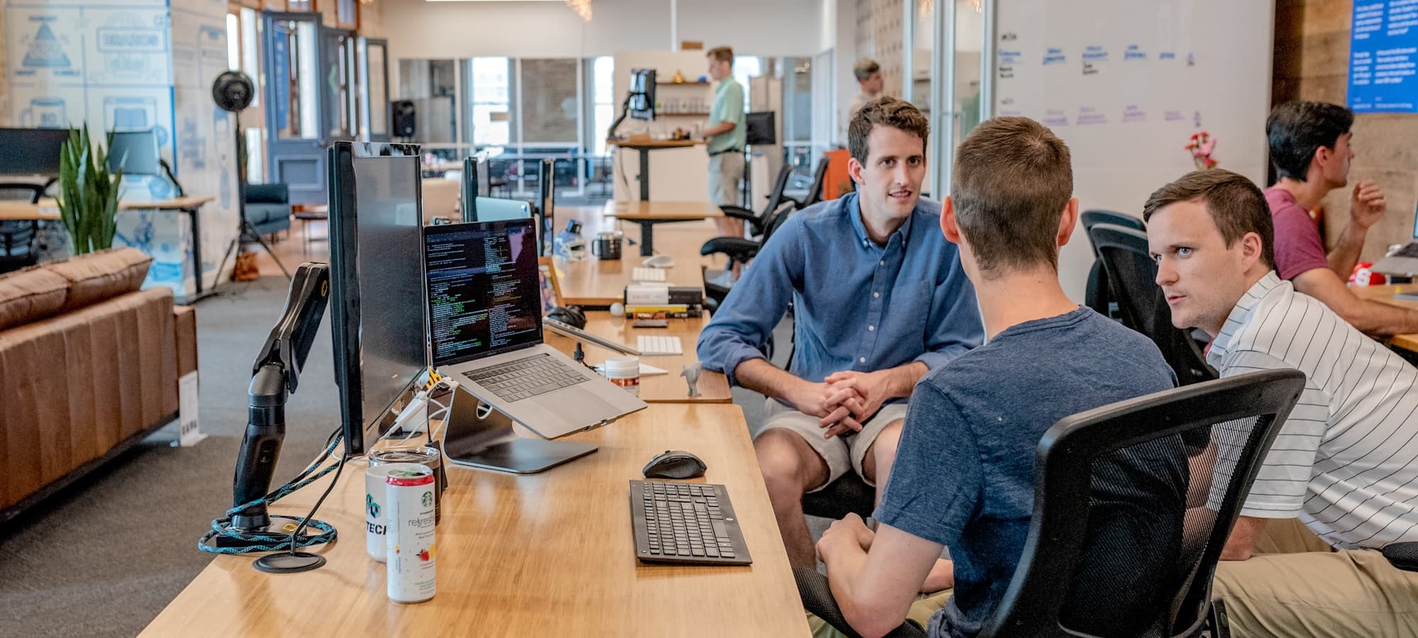 Three colleagues talking at a shared desk in an open plan office, a screen of code open beside them.