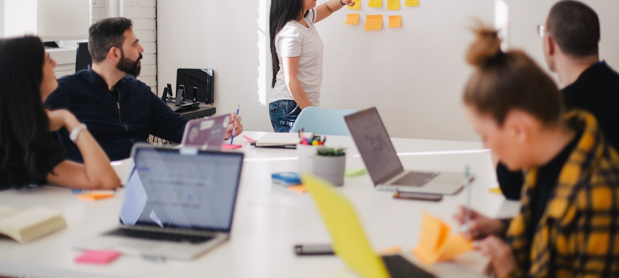 A team seated around a table while a colleague arranges sticky notes on a whiteboard.