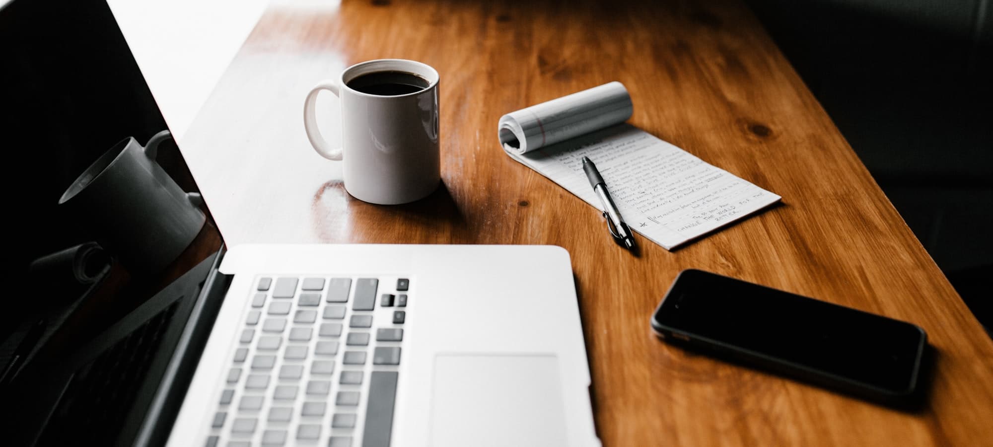 A laptop, a coffee and a notepad of handwritten notes on a wooden desk.